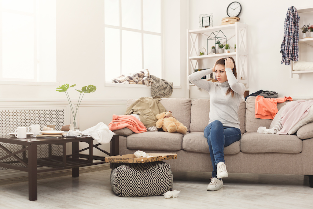 Desperate helpless woman sitting on sofa in messy living room. Young girl surrounded by many stack of clothes. Disorder and mess at home, copy space